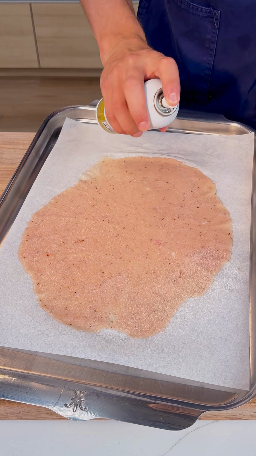 Nick sprays cooking oil over the exposed chicken layer after removing the plastic covering.